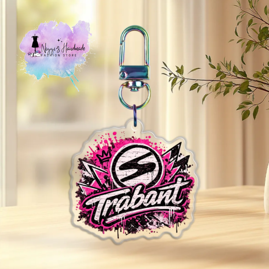 Produktbild eines Trabant-Schlüsselanhängers bei Niggi’s Handmade. Ein Anhänger aus klarem Acryl mit dem Trabant-Markenlogo im pinken und schwarzen Graffiti-Design an einem regenbogenfarben schimmernden Karabinerhaken.