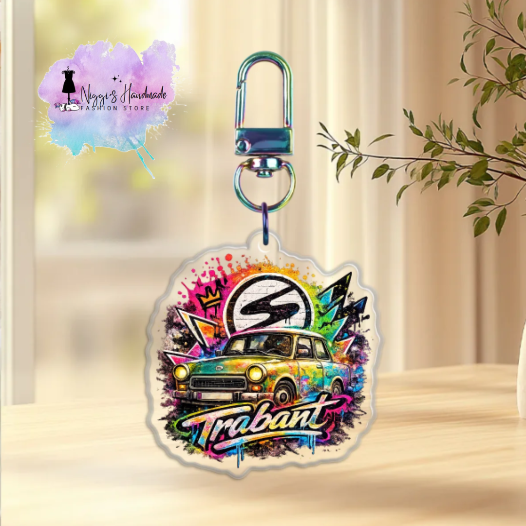 Produktbild eines Trabant-Schlüsselanhängers bei Niggi’s Handmade. Ein runder Anhänger aus klarem Acryl mit einem Trabant 601 und dem Markenlogo in einer bunten, dynamischen Graffiti-Farbexplosion an einem regenbogenfarben schimmernden Karabinerhaken.