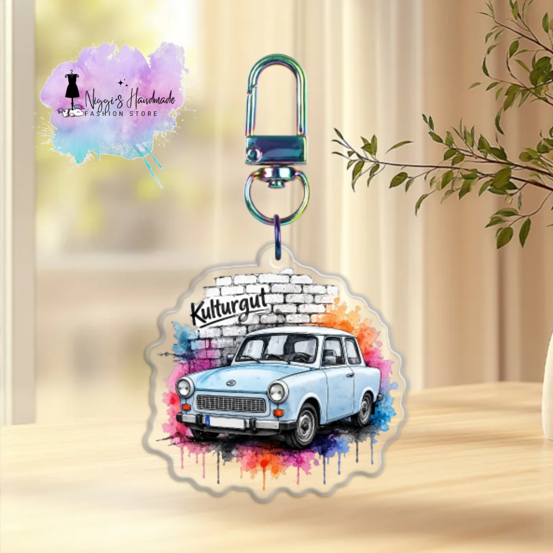 Produktbild eines Trabant-Schlüsselanhängers bei Niggi’s Handmade. Ein runder Anhänger aus klarem Acryl mit dem blauen Aquarell-Trabant 601 und dem Schriftzug 'Kulturgut' an einem regenbogenfarben schimmernden Karabinerhaken.