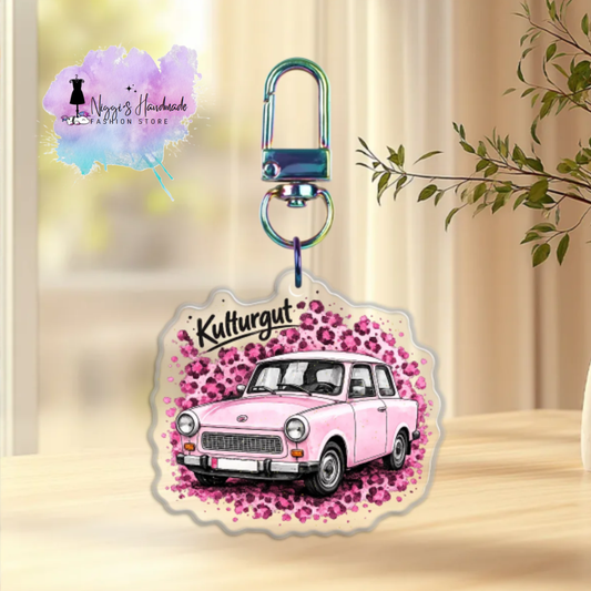Produktbild eines Trabant-Schlüsselanhängers bei Niggi’s Handmade. Ein runder Anhänger aus klarem Acryl mit dem rosa Trabant 601 und dem Schriftzug 'Kulturgut' an einem regenbogenfarben schimmernden Karabinerhaken.