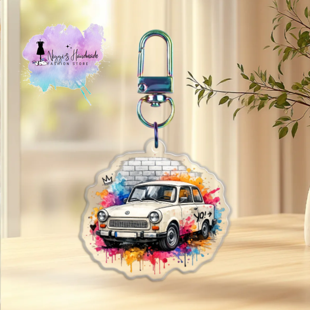 Produktbild eines Trabant-Schlüsselanhängers bei Niggi’s Handmade. Ein runder Anhänger aus klarem Acryl mit einem papyrusweißen Aquarell-Trabant 601 vor einer Steinmauer an einem regenbogenfarben schimmernden Karabinerhaken.