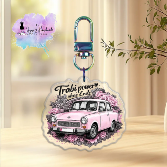 Produktbild eines Trabant-Schlüsselanhängers bei Niggi’s Handmade. Ein Anhänger aus klarem Acryl mit einem rosa Trabant 601 inmitten von rosa Pfingstrosen und dem Schriftzug 'Trabi power ohne Ende' an einem regenbogenfarben schimmernden Karabinerhaken.