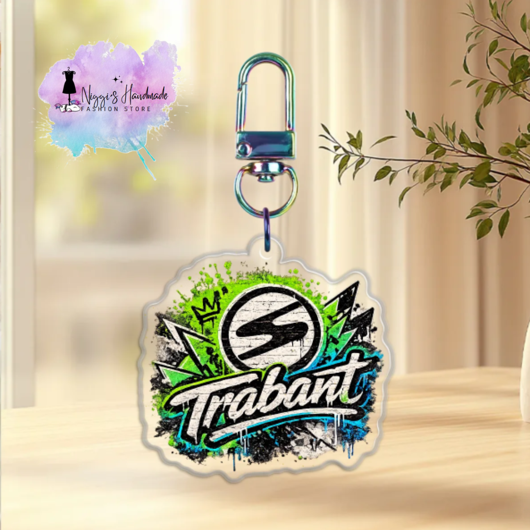 Produktbild eines Trabant-Schlüsselanhängers bei Niggi’s Handmade. Ein Anhänger aus klarem Acryl mit dem Trabant-Markenlogo im neongrünen und blauen Graffiti-Design an einem regenbogenfarben schimmernden Karabinerhaken.