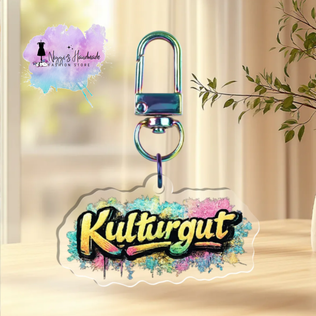Transparenter Acryl-Schlüsselanhänger mit dem Schriftzug „Kulturgut“ im Graffiti-Stil auf buntem Farbklecks-Hintergrund, befestigt an einem rainbow-schimmernden Karabiner.