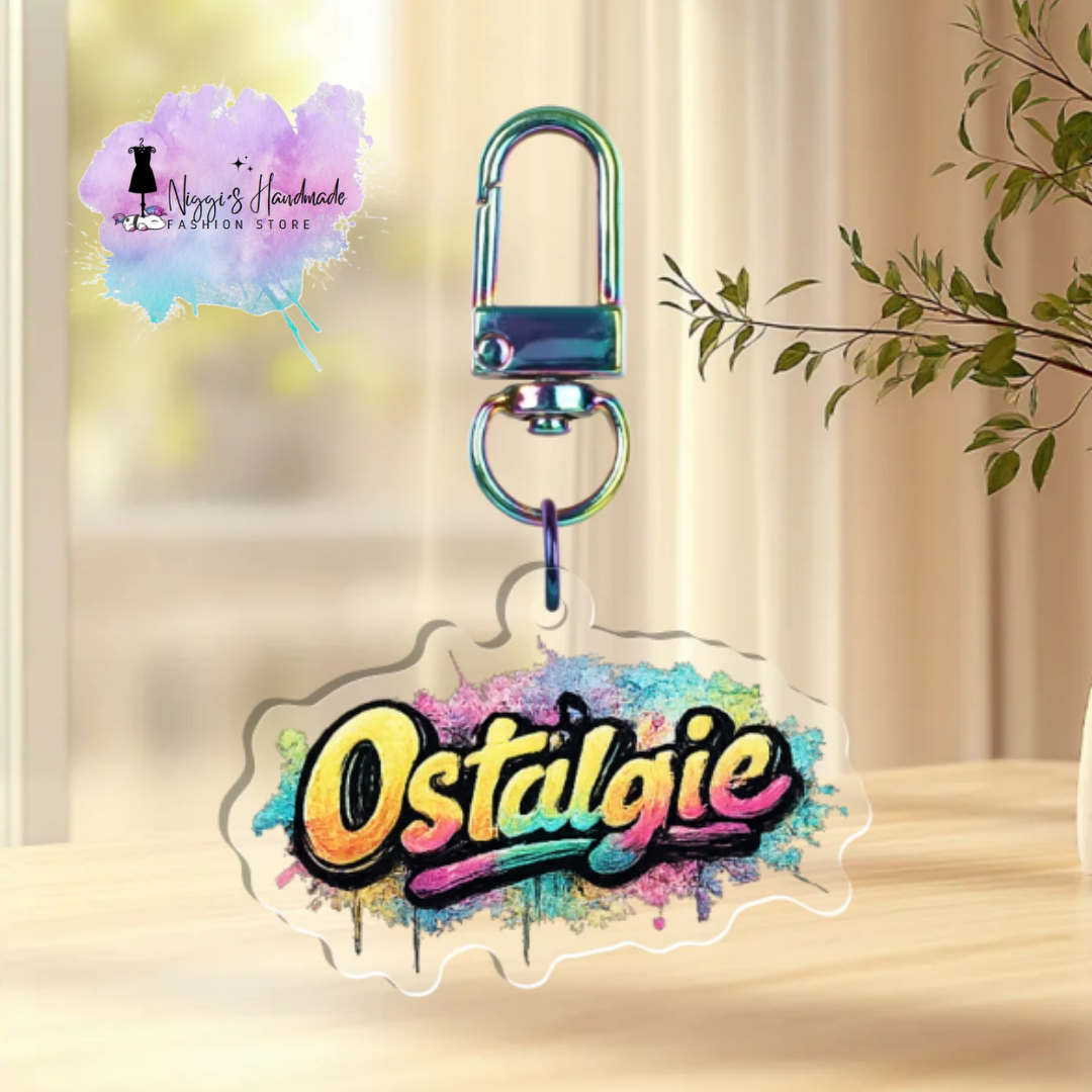 Transparenter Acryl-Schlüsselanhänger mit dem Schriftzug „Ostalgie“ im Graffiti-Stil auf buntem Farbklecks-Hintergrund, befestigt an einem rainbow-schimmernden Karabiner.