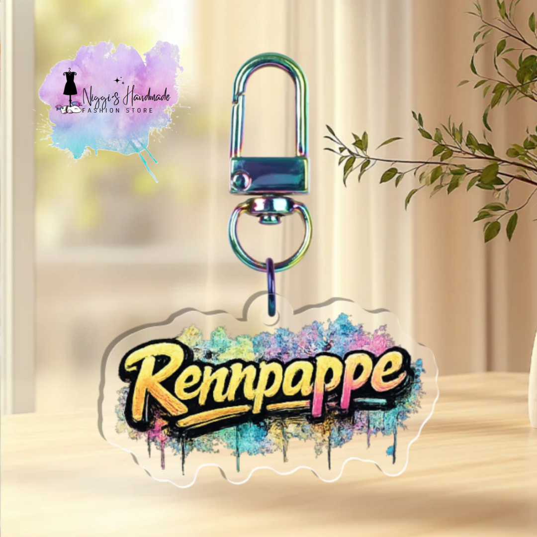 Transparenter Acryl-Schlüsselanhänger mit dem gelb-schwarzen Schriftzug „Rennpappe“ im Graffiti-Look vor bunten Farbspritzern, befestigt an einem metallisch schimmernden Rainbow-Karabiner.