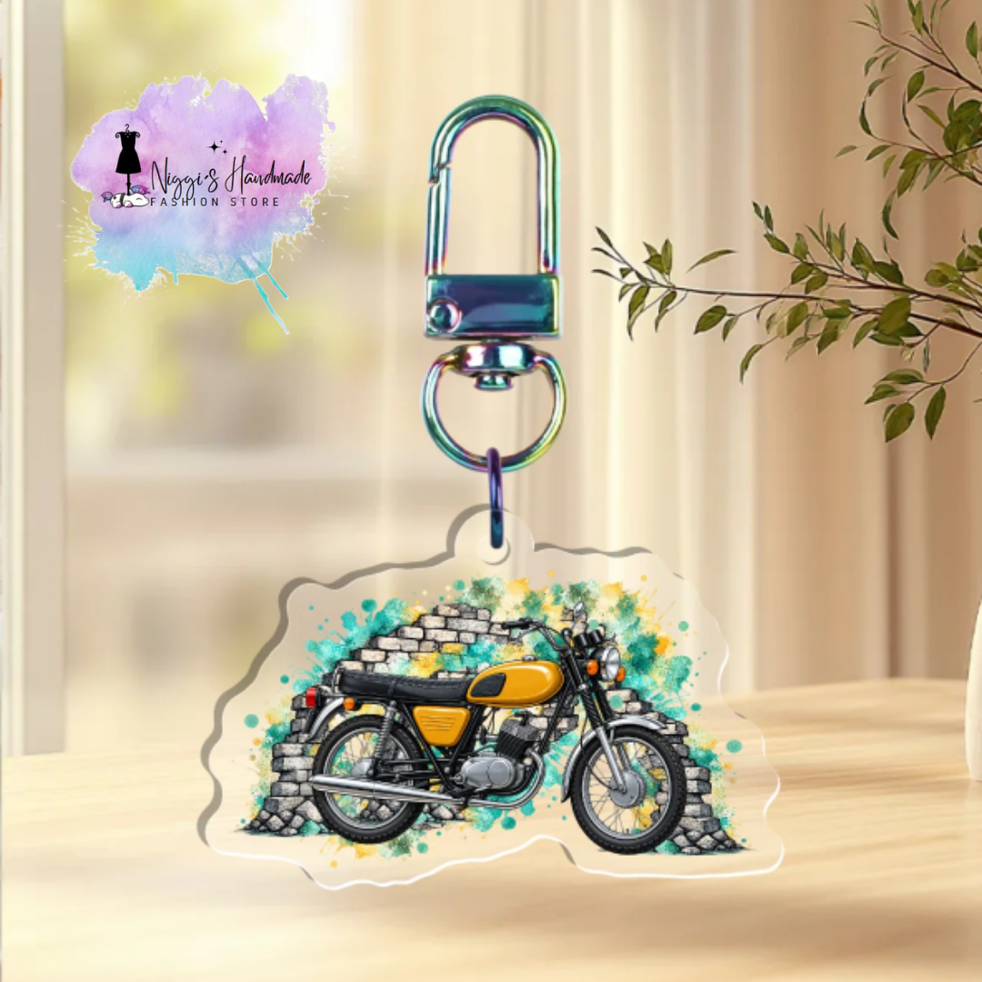 Produktfoto eines runden Schlüsselanhängers aus klarem Acryl mit einem saharabraunen Retro-Moped vor einer weiß-grauen Backsteinmauer und abstrakten Graffiti-Farbspritzern in Türkis und Gelb. Der Anhänger ist an einem metallischen Karabinerhaken mit Rainbow-Effekt befestigt und liegt auf einer hellen Holzoberfläche. Im Hintergrund ist ein zarter grüner Zweig in einer Vase zu sehen. Links oben befindet sich das Logo von „Niggi’s Handmade Fashion Store“.