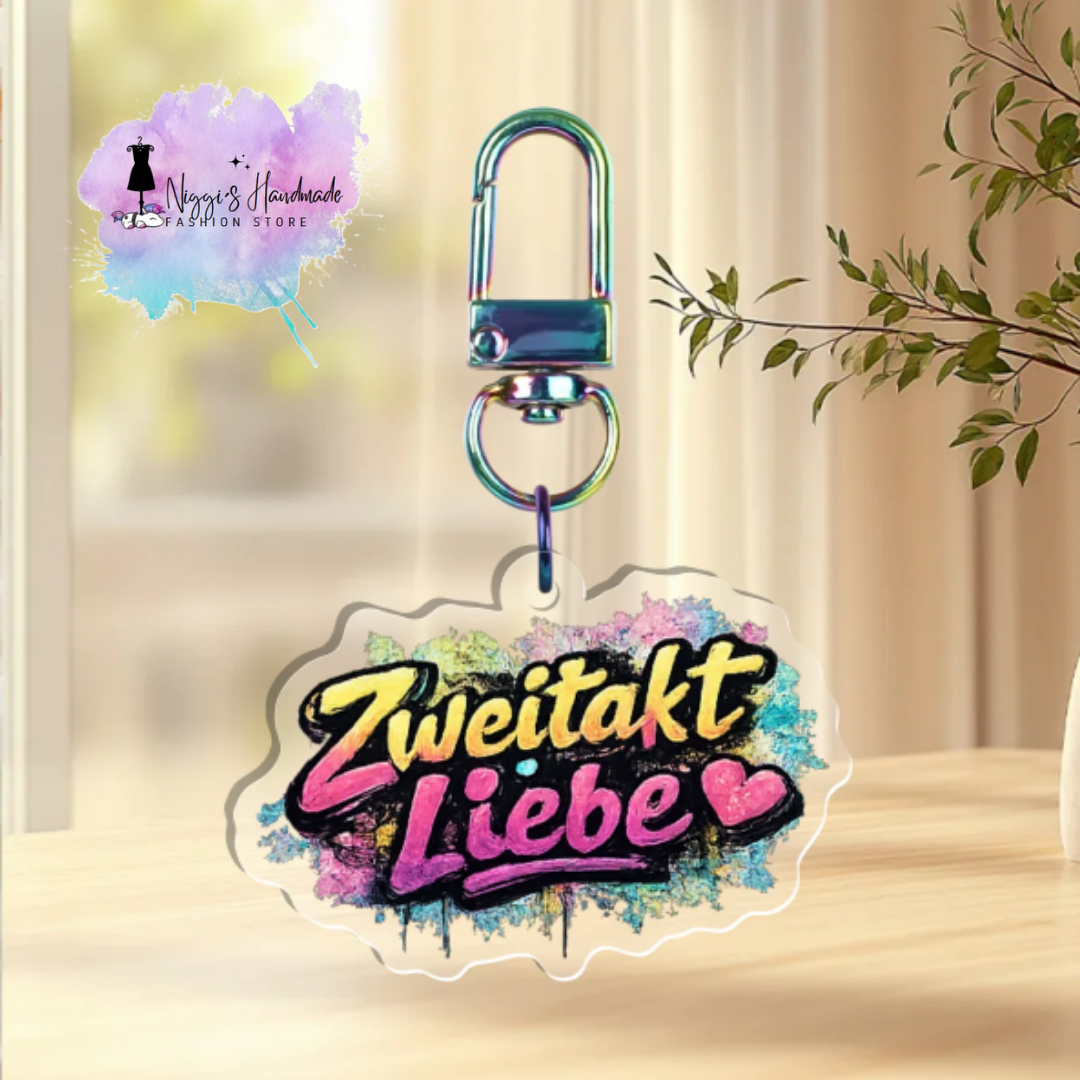 Transparenter Acryl-Schlüsselanhänger mit dem Schriftzug „Zweitakt Liebe“ und zwei Herzen im Graffiti-Stil auf farbenfrohen Farbspritzern, befestigt an einem Rainbow-Karabiner.