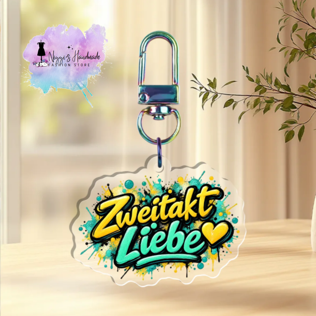 Transparenter Acryl-Schlüsselanhänger mit dem Schriftzug „Zweitakt Liebe“ und zwei gelben Herzen im Graffiti-Stil auf leuchtenden gelb-türkisen Farbspritzern, befestigt an einem Rainbow-Karabiner.