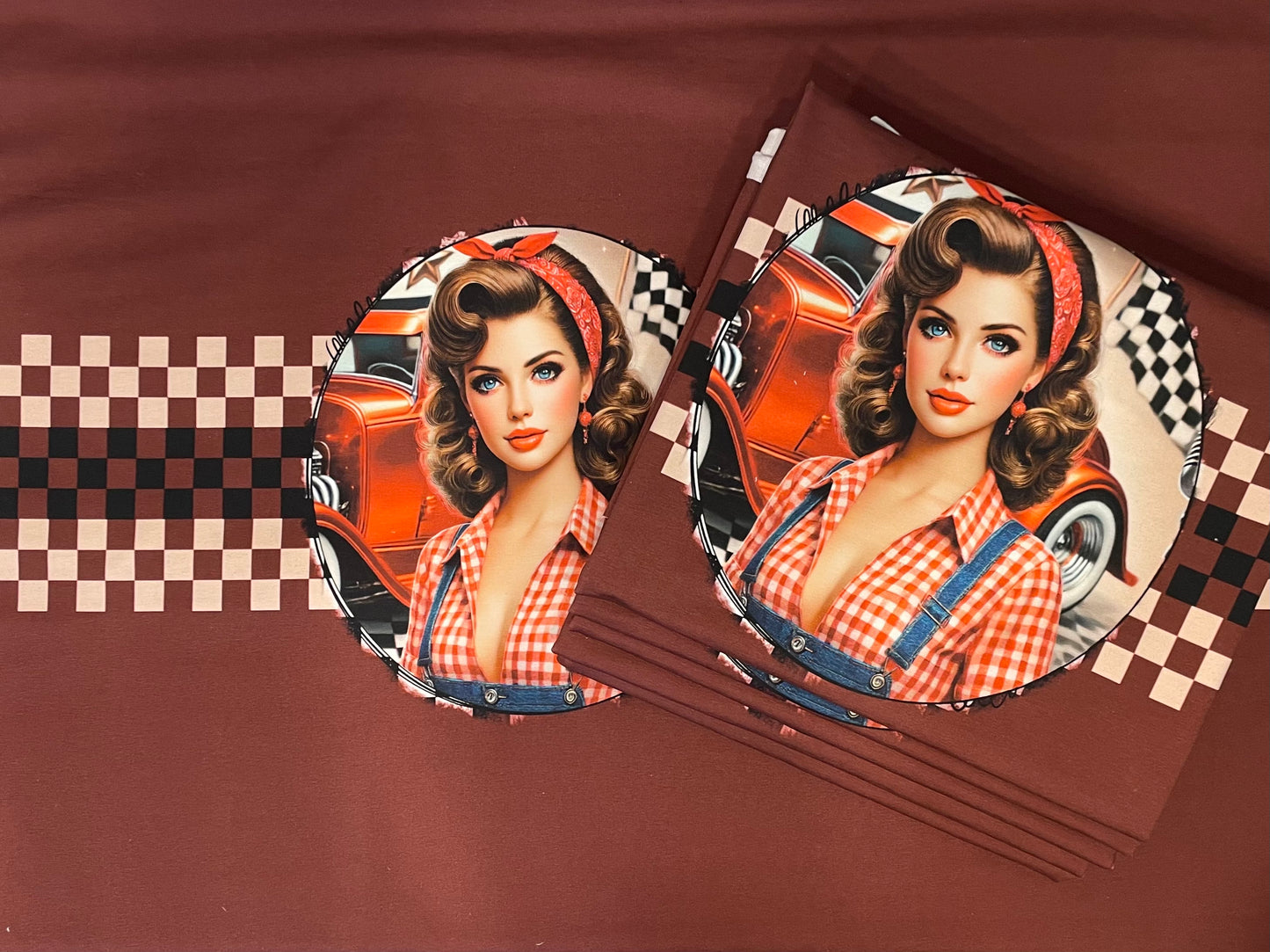 Produktbild des exklusiven French-Terry-Panelstoffes „Vintage Pin-Up Hot Rod“ von Niggi's Handmade Fashion Store. Auf einem kastanienbraunen Stoffhintergrund ist das runde Motiv einer Pin-Up-Frau im 50er-Jahre-Stil mit rot-weiß kariertem Hemd vor einem orangefarbenen Hot-Rod-Oldtimer und einer Zielflagge abgebildet. Rechts daneben liegt ein Stapel desselben gefalteten Stoffes. Ein horizontales, braun-beiges Schachbrettmuster zieht sich über den Stoff.