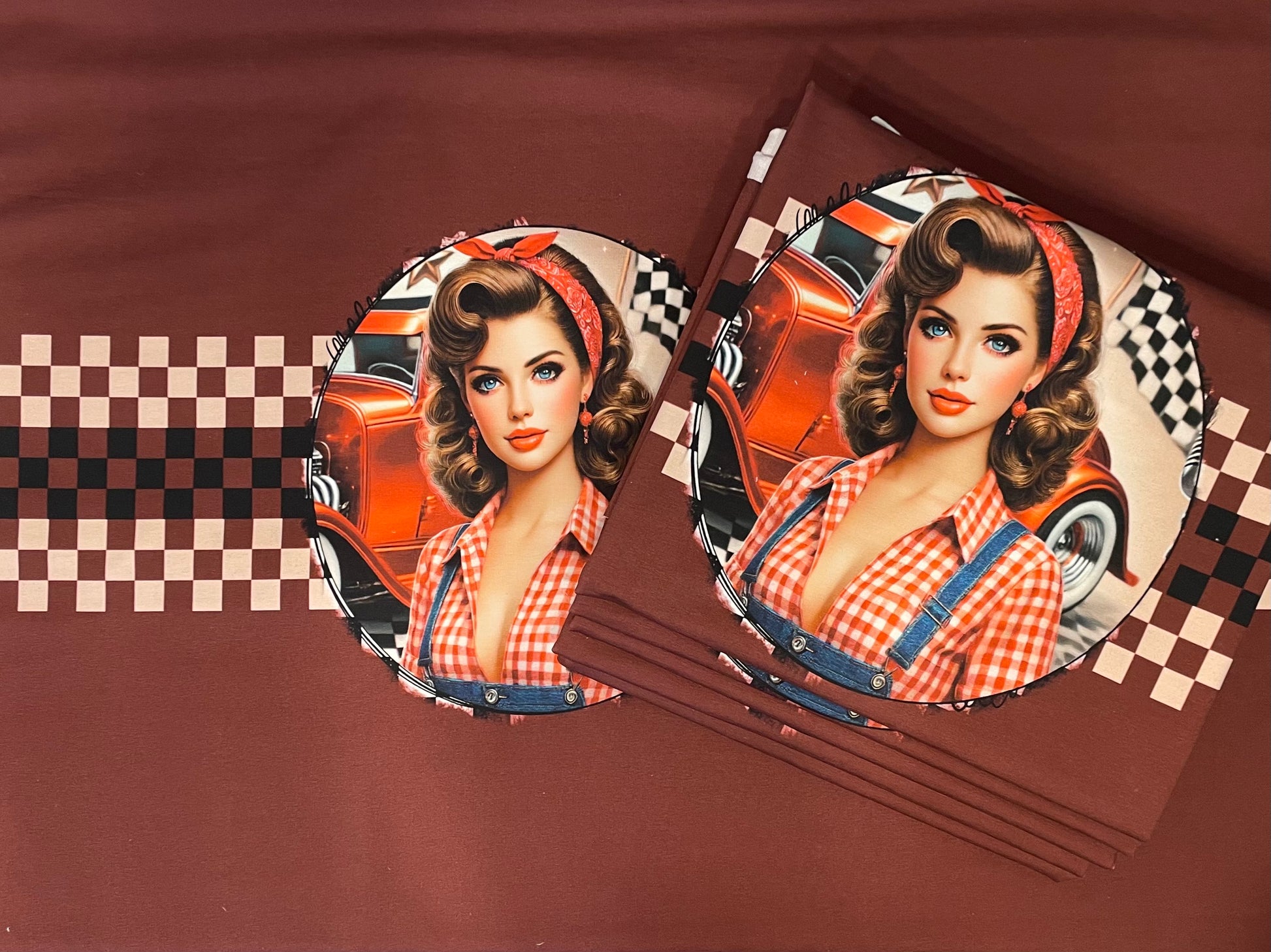 Produktbild des exklusiven French-Terry-Panelstoffes „Vintage Pin-Up Hot Rod“ von Niggi's Handmade Fashion Store. Auf einem kastanienbraunen Stoffhintergrund ist das runde Motiv einer Pin-Up-Frau im 50er-Jahre-Stil mit rot-weiß kariertem Hemd vor einem orangefarbenen Hot-Rod-Oldtimer und einer Zielflagge abgebildet. Rechts daneben liegt ein Stapel desselben gefalteten Stoffes. Ein horizontales, braun-beiges Schachbrettmuster zieht sich über den Stoff.