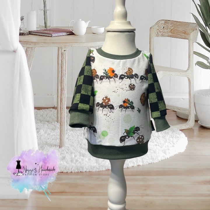 Ein handgemachter Baby-Pullover in Größe 62, präsentiert auf einer hellen Schneiderpuppe. Das weiße Hauptteil zeigt Reihen von Comic-Ameisen, die Blätter, Blumen und Kekse tragen. Die langen Ärmel sind in einem dunkelgrün-schwarzen Schachbrettmuster im Used-Look gestaltet. Der Halsausschnitt sowie die Saum- und Ärmelbündchen sind aus dunkelgrünem Stoff gefertigt. Unten links ist das Logo von Niggi's Handmade Fashion Store abgebildet.