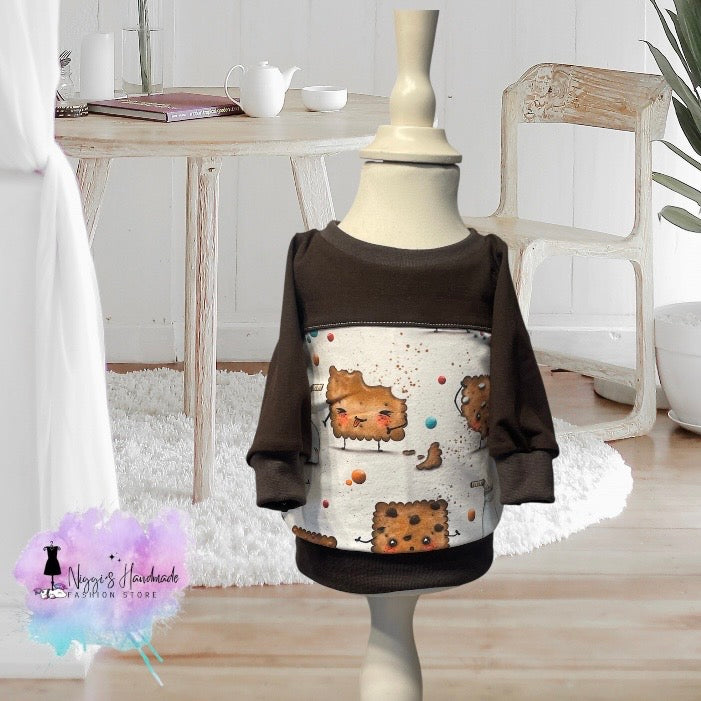 Ein handgemachter Baby-Pullover in Größe 62, präsentiert auf einer hellen Schneiderpuppe. Das helle Hauptteil zeigt lustige Comic-Butterkekse mit Gesichtern, Armen und Beinen inmitten von kleinen bunten Punkten und braunen Krümeln. Die langen Ärmel, der obere Brustbereich sowie alle Bündchen und der Halsausschnitt sind aus dunkelbraunem Stoff gefertigt. Unten links ist das Logo von Niggi's Handmade Fashion Store abgebildet.