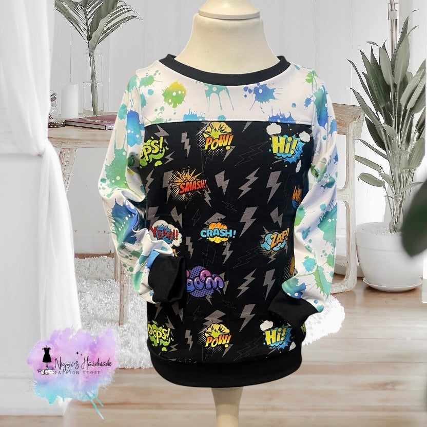 Ein handgemachter Kinder-Pullover in Größe 104, präsentiert auf einer hellen Schneiderpuppe vor einem hellen Wohnzimmer-Hintergrund. Das Oberteil hat ein schwarzes Hauptteil mit Blitzen und bunten Comic-Schriftzügen wie POW und CRASH. Der obere Brustbereich und die Ärmel sind weiß mit neongrünen und blauen Farbspritzern gestaltet. Ausschnitt, Ärmel- und Saumbündchen sind aus schwarzem Stoff gefertigt. Unten links befindet sich das Logo von Niggi's Handmade Fashion Store.