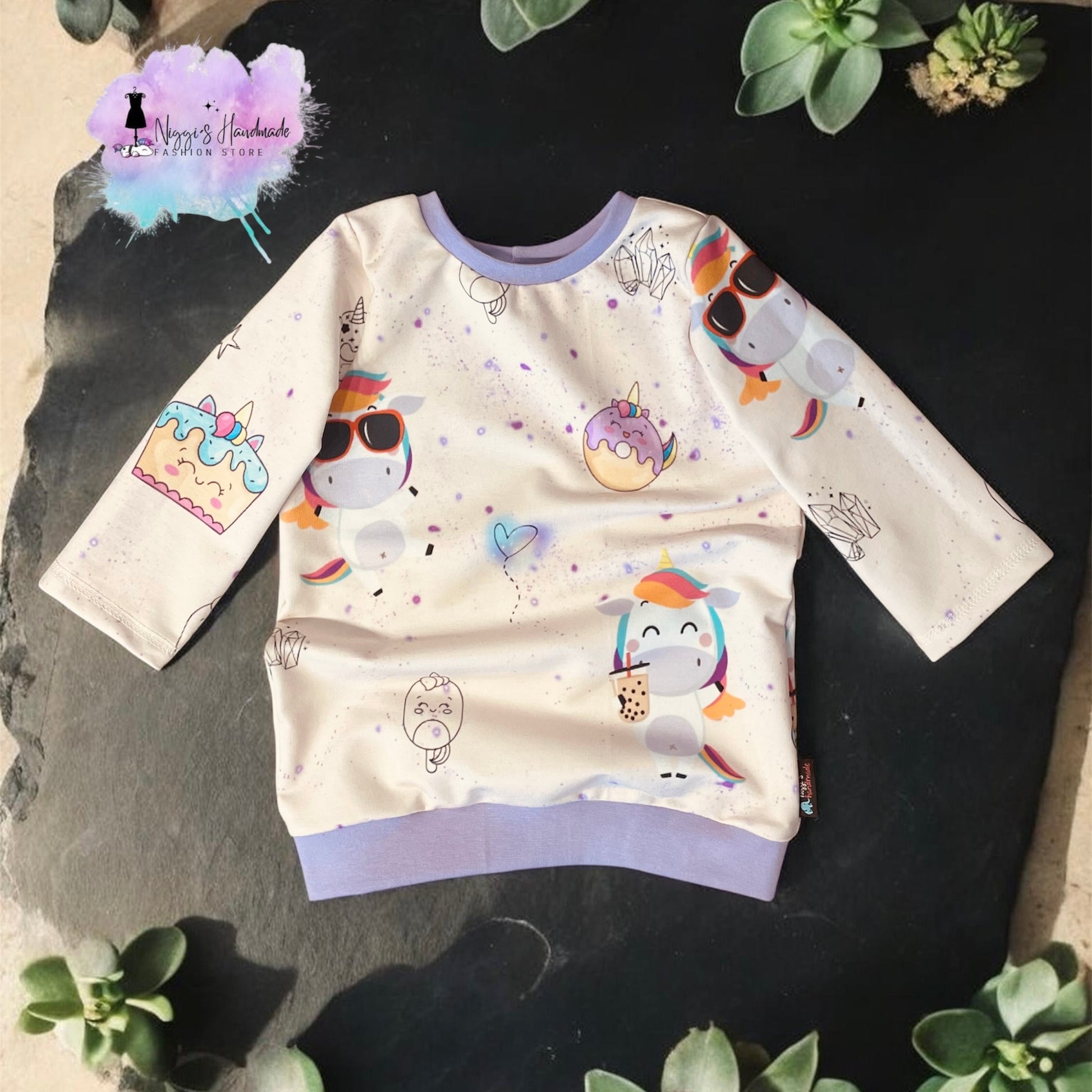 Ein handgemachter Kinder-Pullover aus French Terry in Größe 104, flach liegend auf einer dunklen Schieferplatte präsentiert. Der helle Stoff zeigt bunte Comic-Einhörner mit Sonnenbrillen oder beim Bubble-Tea-Trinken sowie Motive von verzierten Einhorn-Torten und Donuts. Der Halsausschnitt und das breite Saumbündchen sind aus fliederfarbenem Stoff gefertigt. Der Pullover ist umgeben von grünen Sukkulenten-Pflanzen, oben links prangt das Logo von Niggi's Handmade Fashion Store.