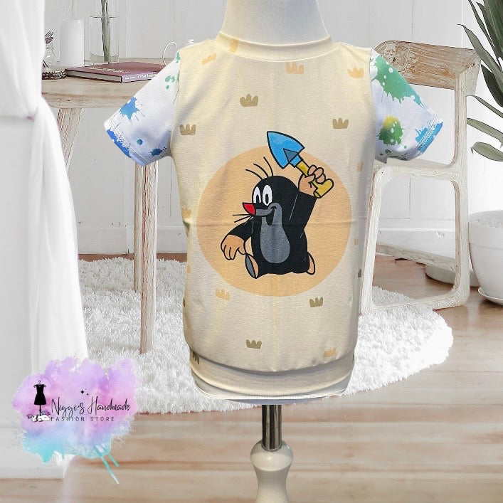 Ein handgemachtes Kinder-T-Shirt mit Pauli dem kleinen Maulwurf in Größe 86, präsentiert auf einer hellen Schneiderpuppe. Das Hauptteil des Kurzarmshirts ist hellbeige mit kleinen Erdhaufen-Mustern und zeigt mittig den kleinen Comic-Maulwurf mit einer blauen Schaufel. Die kurzen Ärmel sind weiß mit auffälligen hellgrünen und blauen Farbspritzern im Graffiti-Look gestaltet. Unten links ist das Logo von Niggi's Handmade Fashion Store abgebildet.