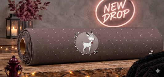 Eine Stoffrolle des French Terry Panels „Winterliche Eleganz“ von Niggi's Handmade liegt auf einem rustikalen Holztisch. Der taupefarbene Stoff zeigt weiße Pünktchen und ein zentrales Motiv: Ein schlafendes, weiß-graues Rehkitz mit einer roten Schleife, umrahmt von einem filigranen Blätterkranz. Im Hintergrund leuchtet ein neonfarbenes „New Drop“-Schild.
